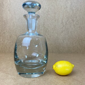 Puede incluir: Una decantador de vidrio transparente con un tap&oacute;n de vidrio azul. La decantador tiene un cuerpo redondeado y una base ancha. Un lim&oacute;n amarillo se encuentra al lado de la decantador.