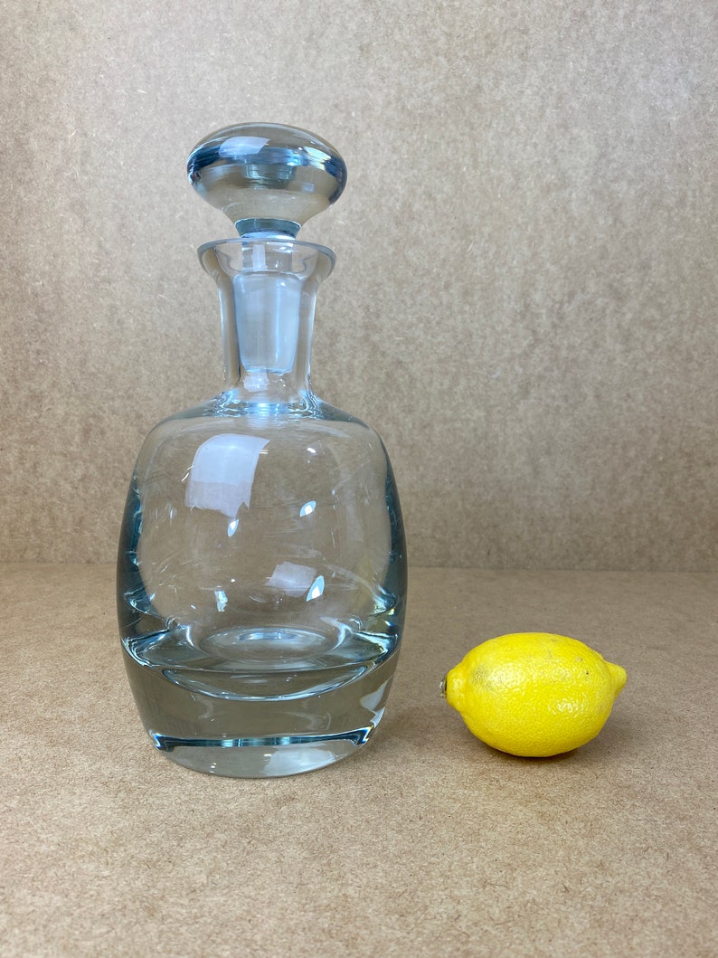 Puede incluir: Una decantador de vidrio transparente con un tap&oacute;n de vidrio azul. La decantador tiene un cuerpo redondeado y una base ancha. Un lim&oacute;n amarillo se encuentra al lado de la decantador.
