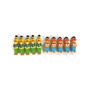 Op de afbeelding: Een verzameling kleine, kleurrijke beeldjes. De beeldjes hebben de vorm van een stripfiguur met een gele huid, zwart haar en een rode neus. Ze dragen verschillende outfits, waaronder groen-blauwe gestreepte shirts en een blauwe jeans.