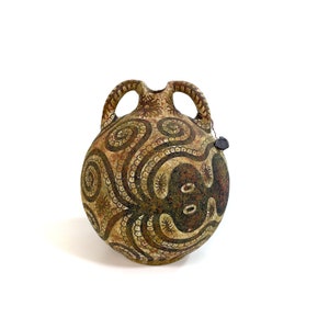 May include: A round, antique-style ceramic jug with two handles and a spout. The jug features a brown and beige color scheme with intricate spiral and dot patterns. A black seal hangs from the jug.