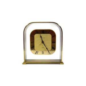 May include: A gold-toned quartz desk clock with a square face and Roman numerals. The clock face is a light gold color with black hands and the word "SERENA" printed on the face. The clock is encased in a clear, arched frame.