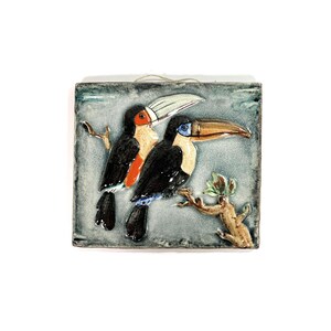 Può includere: Una piastrella di ceramica con un design 3D di due tucani appollaiati su un ramo. I tucani sono dipinti in tonalità di nero, bianco, arancione e blu. Lo sfondo è di colore grigio-azzurro chiaro.