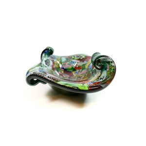 Könnte beinhalten: Eine farbenfrohe, handgefertigte Glasschale mit einem einzigartigen Design. Die Schale weist eine Mischung aus Farben auf, darunter Grün, Rot und Blau, mit einem Wirbelmuster. Sie hat zwei geschwungene Griffe. Die Schale hat einen Durchmesser von etwa 15 cm.