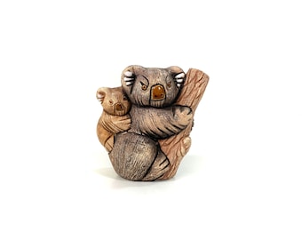 May include: A ceramic koala figurine depicting a mother and baby koala on a tree branch. The koalas are brown and tan with detailed fur and a glossy finish. The figurine is set against a white background. The figurine is a decorative item.