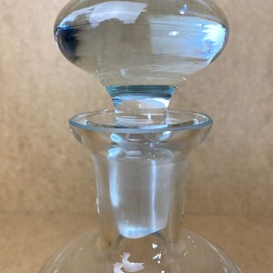 Puede incluir: Una botella de vidrio transparente con un tap&oacute;n redondo de vidrio transparente. La botella tiene una base ancha y un cuello estrecho.