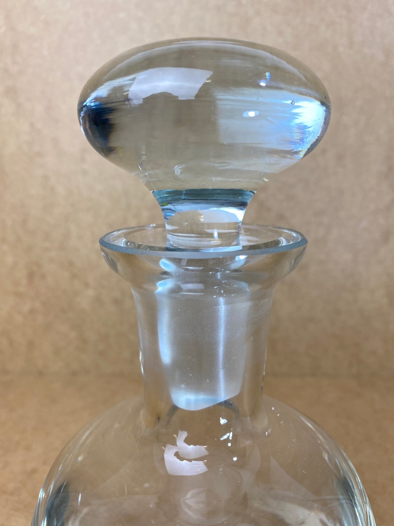 Puede incluir: Una botella de vidrio transparente con un tap&oacute;n redondo de vidrio transparente. La botella tiene una base ancha y un cuello estrecho.