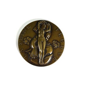 May include: Bronze round relief sculpture featuring a nude female figure holding a draped cloth, with a ship and grapes. The text "LA SEINE FLEUVE" is inscribed around the edge. The sculpture has a detailed, antique appearance.