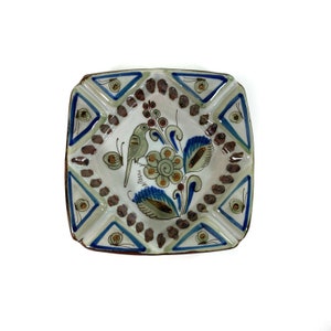May include: Square ceramic ashtray with a blue, green, and gold floral design and a bird. The ashtray has a brown rim and a white background.