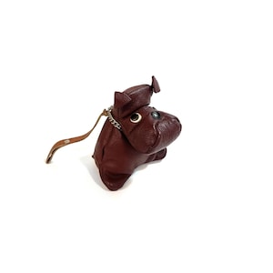 May include: A burgundy leather coin purse in the shape of a hippo. The hippo has small, round, gold-coloured eyes and a silver chain attached to a leather strap. The purse is on a white background.