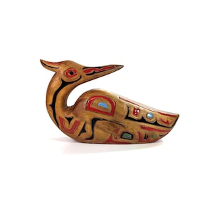 May include: A hand-carved wooden bird figurine featuring a natural light brown wood grain finish. The surface is decorated with stylized black, red, and light blue painted geometric patterns that follow the contours of the bird's body.