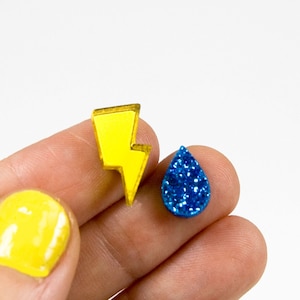 Può includere: Un paio di orecchini, uno a forma di fulmine giallo e l'altro a forma di goccia blu con glitter.