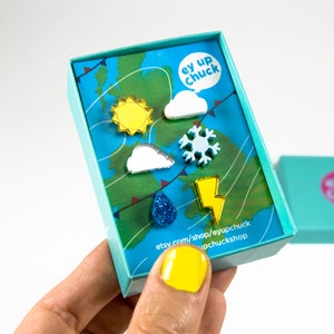Può includere: Un set di sei orecchini a tema meteorologico in una scatola blu. Gli orecchini sono in acrilico e presentano un sole, una nuvola, un fiocco di neve, un fulmine, una goccia di pioggia e un'altra nuvola. La scatola ha il testo "ey up chuck" su di essa.