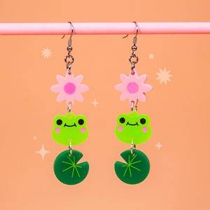 Peut inclure: Boucles d'oreilles pendantes avec un motif de marguerite, de grenouille verte et de nénuphar. Les boucles d'oreilles ont des crochets argentés et sont suspendues à une barre rose sur un fond pêche. La grenouille a des yeux noirs et des joues roses.