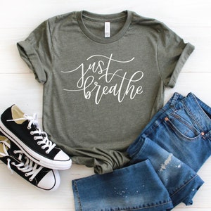 May include: A green t-shirt with the text "just breathe" in white cursive lettering. The shirt is laid flat on a white surface with a pair of black sneakers and blue jeans.