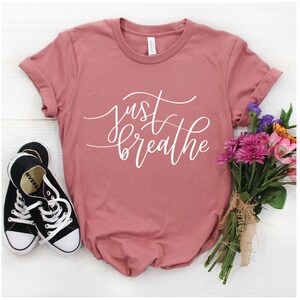 May include: A dusty rose t-shirt with the text "just breathe" in white script lettering. The shirt is photographed on a white surface with a pair of black sneakers and a bouquet of pink and purple flowers.
