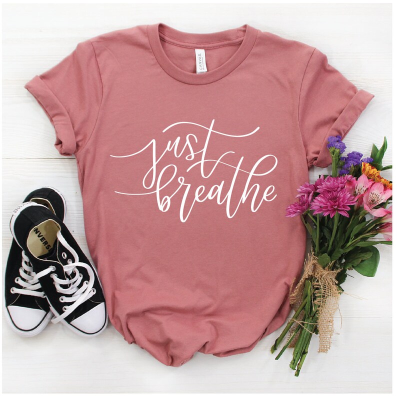 May include: A dusty rose t-shirt with the text "just breathe" in white script lettering. The shirt is photographed on a white surface with a pair of black sneakers and a bouquet of pink and purple flowers.