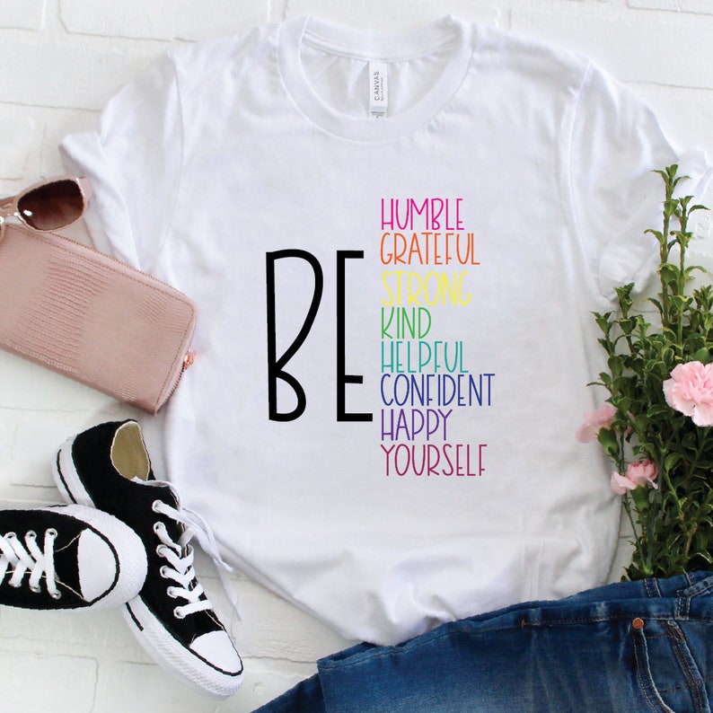 Puede incluir: Camiseta blanca con la palabra "BE" en negro y una lista de atributos positivos en colores arco&iacute;ris: humilde, agradecido, fuerte, amable, servicial, seguro, feliz, t&uacute; mismo.