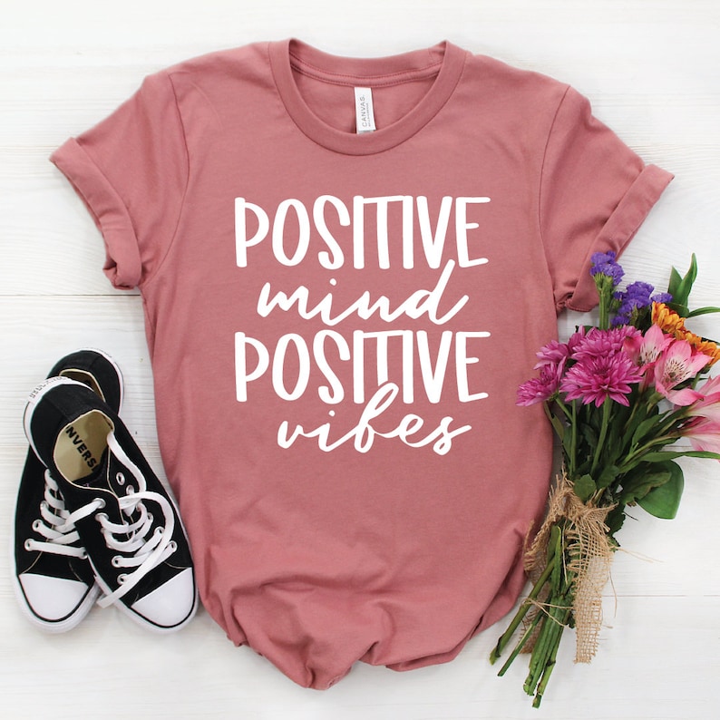 May include: A dusty rose t-shirt with the text "POSITIVE mind POSITIVE vibes" in white lettering. The shirt is laid flat on a white wooden surface with a pair of black sneakers and a bouquet of pink and purple flowers.