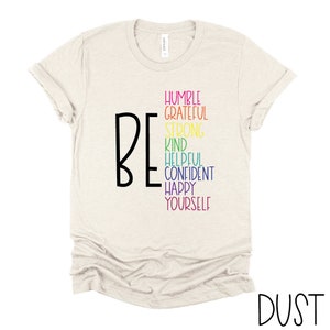 Puede incluir: Una camiseta beige con la palabra "BE" en negro y una lista de atributos positivos en colores arco&iacute;ris: humilde, agradecido, fuerte, amable, servicial, seguro, feliz, t&uacute; mismo.