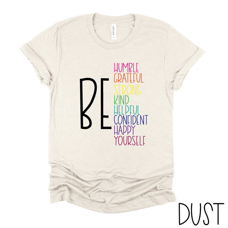 Puede incluir: Una camiseta beige con la palabra "BE" en negro y una lista de atributos positivos en colores arco&iacute;ris: humilde, agradecido, fuerte, amable, servicial, seguro, feliz, t&uacute; mismo.
