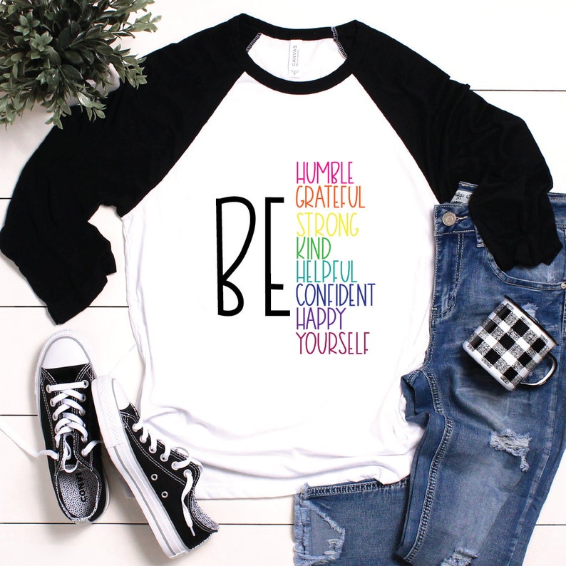Puede incluir: Una camiseta de b&eacute;isbol de manga larga blanca con mangas negras. La camiseta tiene la palabra "BE" en negro y una lista de palabras positivas de colores arco&iacute;ris: "HUMBLE, GRATEFUL, STRONG, KIND, HELPFUL, CONFIDENT, HAPPY, YOURSELF".