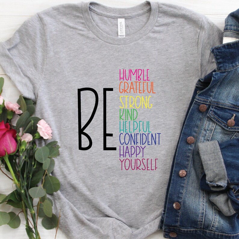 Puede incluir: Camiseta gris con la palabra "BE" en negro y una lista de atributos positivos en colores arco&iacute;ris: Humilde, Agradecido, Fuerte, Amable, Servicial, Seguro, Feliz, T&uacute; mismo.