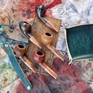 Pode incluir: Cinco cachimbos de madeira em uma bandeja de madeira com uma bolsa de couro verde. Os cachimbos são de vários tons de marrom e têm um acabamento natural. A bolsa tem a palavra "Briar" gravada em relevo.