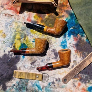 Puede incluir: Tres pipas de fumar de madera con tallos de diferentes colores. Las pipas están sobre un fondo de tela colorido.