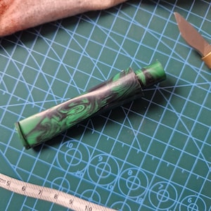 May include: A green and black marbled pen cap with a swirling pattern. The cap is made of a hard plastic material and is designed to fit a pen.