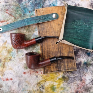 May include: Two tobacco pipes with black stems and brown bowls rest on a wooden surface. A teal leather tray and wristband with the name "Dalandis" are also present. The background has a colorful, abstract pattern.
