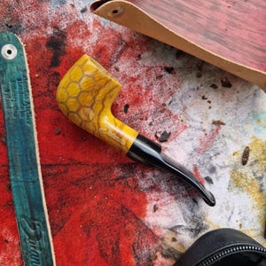 May include: A yellow tobacco pipe featuring a carved honeycomb pattern on the bowl and the word Salantis engraved on the side. The pipe has a smooth, black acrylic stem and a polished finish, showcasing the natural wood grain of the bowl.