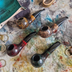Pode incluir: Quatro cachimbos de tabaco artesanais com bocais curvos e fornilhos arredondados são exibidos. Os cachimbos apresentam uma variedade de cores e padrões, incluindo castanho, preto, vermelho e verde. Os fornilhos têm acabamentos texturizados e os bocais são lisos e brilhantes.