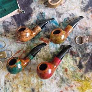 Puede incluir: Cuatro pipas de madera con tallos negros y manchas de diferentes colores. Las pipas están dispuestas sobre una superficie moteada y colorida.
