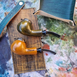 Puede incluir: Dos pipas de tabaco de madera, una verde oliva y otra marrón claro, descansan sobre una bandeja de madera. Cada pipa tiene una boquilla negra y una tapa de metal. También se ven una bandeja y una correa de cuero verde azulado.