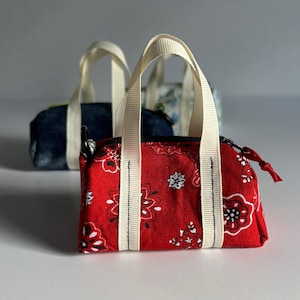 May include: Three small fabric handbags with white handles are displayed. The frontmost bag is red with a white paisley pattern. The other two bags are blue and floral patterned. Each bag has a zipper closure.