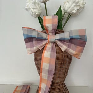 May include: A woven wicker vase with a large, colorful plaid bow. The bow features shades of orange, purple, and blue. White tulip flowers with green stems and leaves are inside the vase. The bow's ribbon hangs down the side.