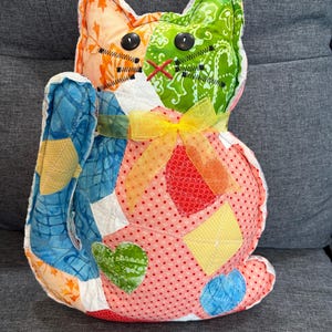 May include: A handmade patchwork cat-shaped pillow. The cat features a colorful design with orange, green, blue, and red fabrics. It has black button eyes, stitched details, and a yellow ribbon bow. The pillow is a unique decorative item.