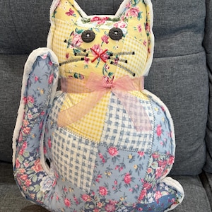 May include: A handmade cat-shaped pillow with a patchwork design. The cat features a yellow floral face, button eyes, and stitched whiskers. The body combines floral, gingham, and solid fabrics in blue, yellow, and white, with a pink ribbon bow.