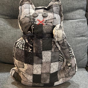May include: A stuffed cat toy constructed from a patchwork of various black, white, and gray patterned fabrics. The cat features two dark button eyes, a stitched red "X" for a nose, and a black ribbon tied in a bow around its neck.