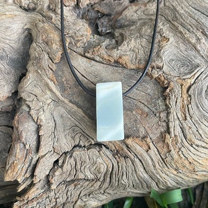 May include: A rectangular, pale blue-green stone pendant suspended from a thin, black cord. The pendant has a smooth, polished surface with subtle, light-colored banding, and it hangs vertically from the simple, dark, circular necklace cord.