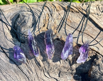 Könnte beinhalten: Sechs Amethyst-Kristallketten mit schwarzen Kordeln. Die rohen, lila Kristalle variieren in Form und Größe, jede ist an einer dünnen, schwarzen Kordel befestigt. Die Halsketten werden auf einem verwitterten, strukturierten Holzstück mit grünem Laub im Hintergrund präsentiert.