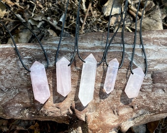 May include: Five crystal pendant necklaces with black cords are displayed on a weathered wooden log. The crystals vary in size and color, ranging from clear to pale pink. The necklaces are arranged in a row, showcasing their polished, pointed shapes.