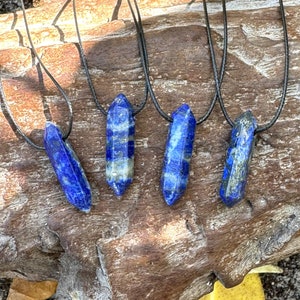 May include: Four blue lapis lazuli crystal necklaces with black cords. The crystals are a deep blue color with streaks of lighter blue and gray. The necklaces are displayed on a piece of weathered wood.