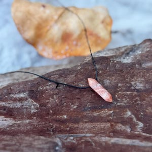 Pode incluir: Colar com pingente de pedra natural. O pingente é de cor rosa-alaranjado claro, com formato geométrico. O colar tem um cordão preto. O pingente é exibido em um pedaço de madeira.