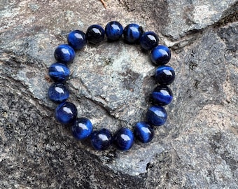 Puede incluir: Un brazalete de cuentas de ojo de tigre azul. El brazalete está hecho de cuentas redondas y pulidas con un color azul oscuro y un efecto reflectante de ojo de gato. Descansa sobre una superficie rocosa gris texturizada.