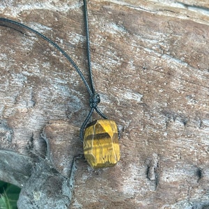 Puede incluir: Collar con colgante de ojo de tigre amarillo dorado facetado con rayas marrones. La piedra está suspendida de un cordón negro. El collar se muestra sobre una superficie de madera envejecida.