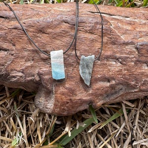May include: Two necklaces with natural stone pendants. One pendant is rectangular with blue and white hues, the other is a gray, irregularly shaped stone. Both are suspended on black cords, displayed on a brown wooden log.