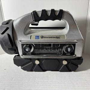 May include: A silver and black Stormtracker by VTech weather radio and flashlight combination device. The unit features a top handle, a front-facing radio dial with volume and tuning knobs, and a large circular flashlight head on the left side.