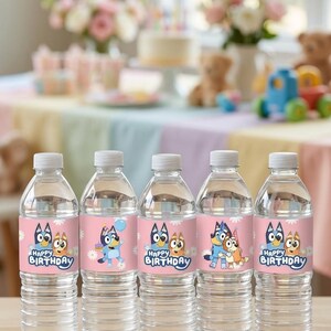 May include: Five clear plastic water bottles featuring pink labels with white daisy patterns and illustrations of Bluey and Bingo characters. The labels include the text Happy Birthday in blue lettering, with various poses of the characters.