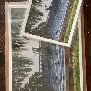 Pu&ograve; includere: Un dipinto ad acquerello incorniciato che rappresenta una scena di lago con una piccola struttura in legno sul bordo dell'acqua. L'acqua &egrave; di un blu profondo con increspature bianche. La struttura &egrave; circondata da alberi e erba verdi.